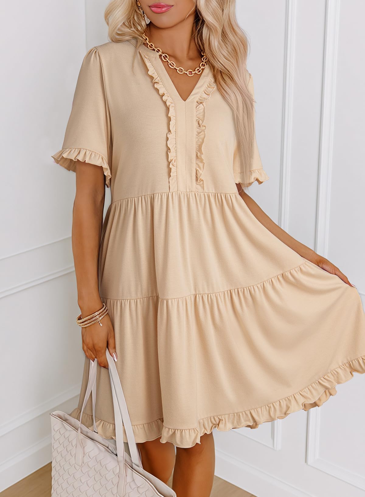 Woman wearing a beige dress with ruffled details, holding a white bag against a white wall.